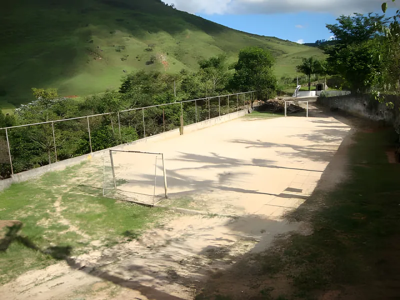 Foto real: campo de areia com traves de futebol, cercado de mato, na Colônia